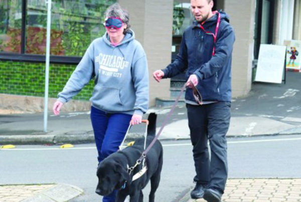 woman wearing a blindfold walking with a Guide Dog and man