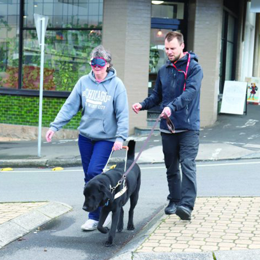 woman wearing a blindfold walking with a Guide Dog and man