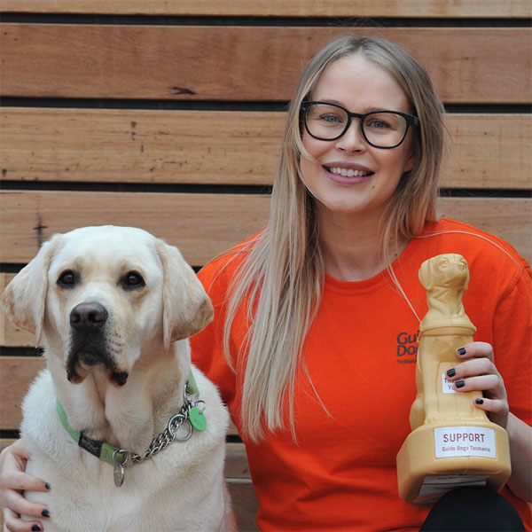 Woman in orange t-shirt sits next to yellow labrador, while holding a Mini Dog collection box