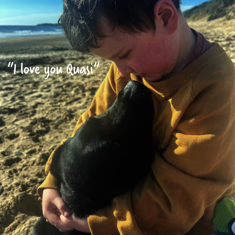 A young boy on the beach cuddling his black dog and looking into his eyes with the text "I love you Quasi".