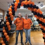 2 Volunteers stand under a balloon arch, smiling.