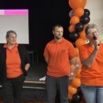 Three people in orange shirts stand in front of a stage, with one speaking into a mic.