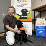 A man kneels beside a black labrador in an orange Guide Dog in Training vest.