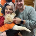 A woman dressed as Cruella de Vil poses holding a Guide Dog in Training in her arms. A man stands with her.